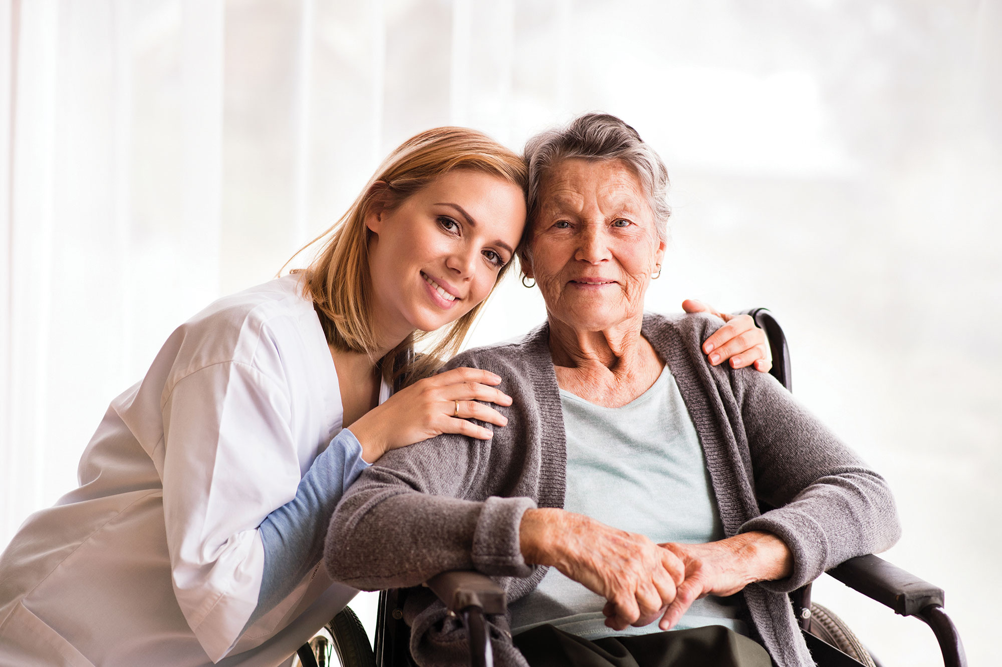 一个金发碧眼的女人穿着白色护理外套跪在一个老妇旁边，坐在轮椅上的白发短。他们俩都在微笑。