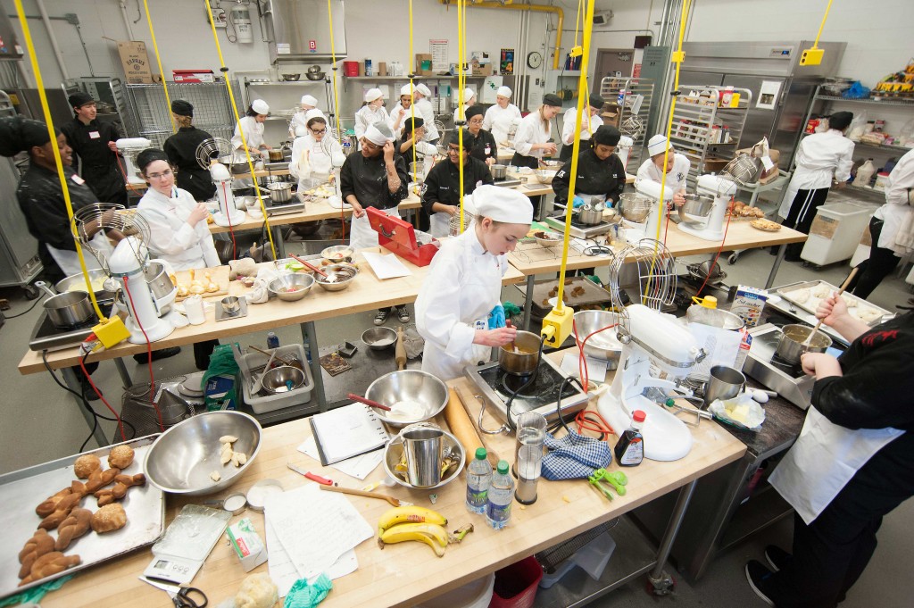 许多穿着厨师制服的学生在烘焙实验室里忙着烹饪。