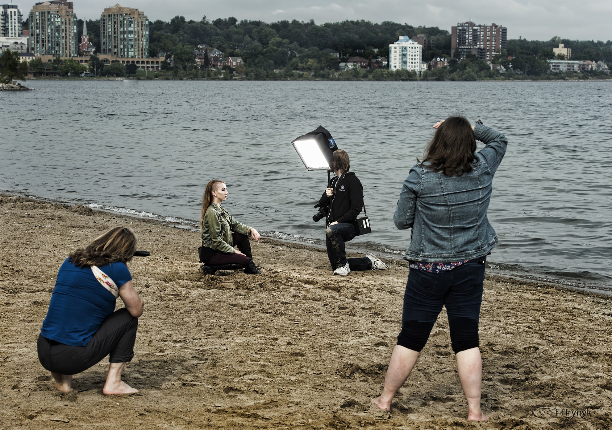 一名女性跪在沙滩上，在两名女摄影师面前摆姿势，另一名女性拿着软盒照明设备